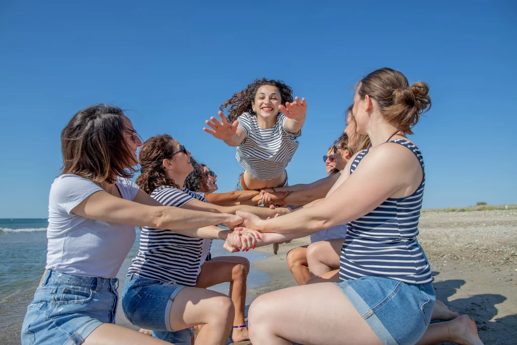 Photographie faite lors d'une séance photo evjf à la plage de Carnon, les filles sont à genoux mains croisées et la mariée au centre saute les mains en avant, avec un grand sourire. Photographie faite par un photographe evjf à Montpellier