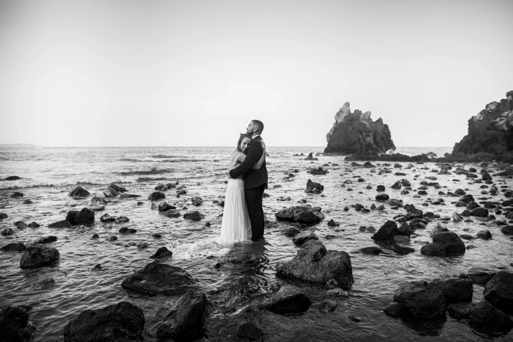 Photographie de mariage, Mathilde et Guillaume, photo à la plage de la gradn conque. Ils sont au millieu des rochers dans l'eau et se fond un calin, on peut voir en arrière plan le grand rocher et les petits rochers en premier plan. Photo en noir et blanc. Photographie prise par un photographe de mariage professionnel
