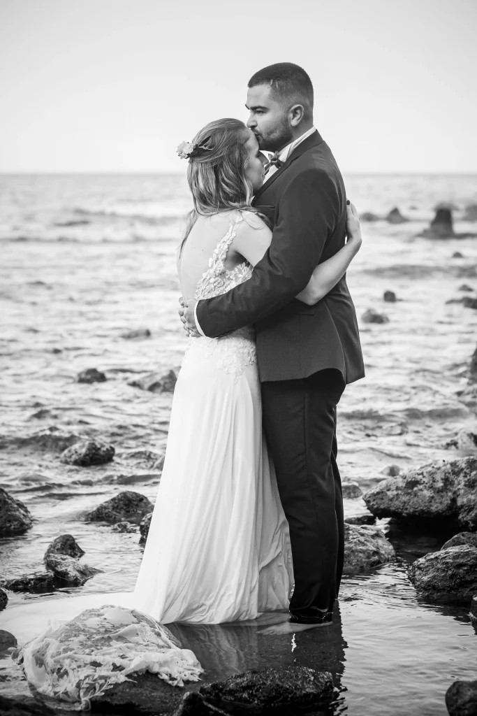 couple de marié durant la séance de couple qui se font un câlin en face à face à la plage parmis les rochers dans l'eau, photographier par un photographe de mariage.