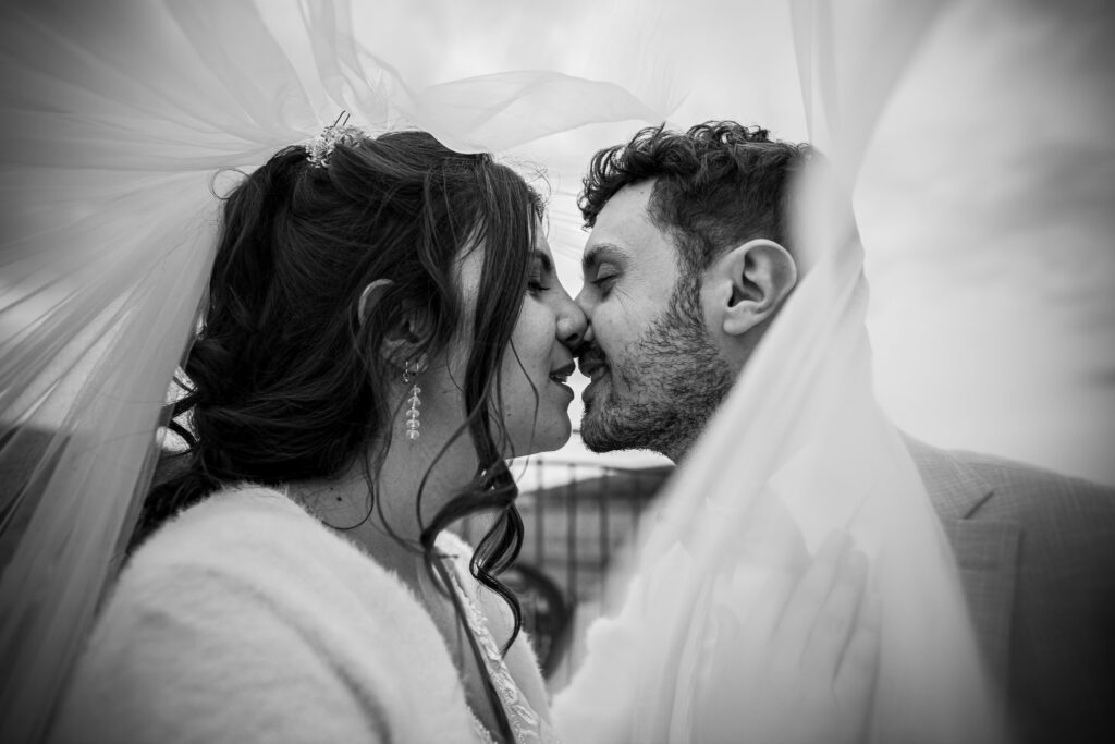 photographie de mariage Ambre et Léo. Photo de couple en noir et blanc. Les deux mariés s'embrasse sour le voile, photo prise au 24 mm avec un effet de proximité et du voile qui fait un ligne directrice vers les deus amoureux. Photographie prise par un photographe de mariage professionnel à Nîmes