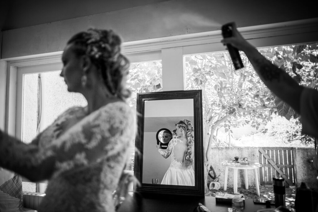 Mariage de Audrey et John dans l'hérault, la mariée se prépare, la coiffeur passe un dernier coup de laque sur les cheveux de la mariée qui se regarde dans un miroir et qui est photographier de dos fasse à un autre miroir qui laisse apparaitre le visage de la mariée