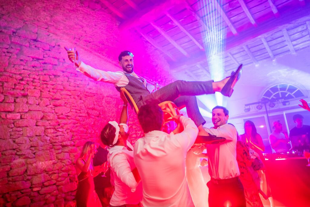 Photo de Mariage de Maxime et Muge en contreplongée . Un invité qui est projeter en l'air par deux autres invit&s, il le tiennent par les jambes et lui à les bras lever vers le ciel. Il porte une chemise blanche et un pantalon beige. Photographie prise par un photographe de mariage professionnel