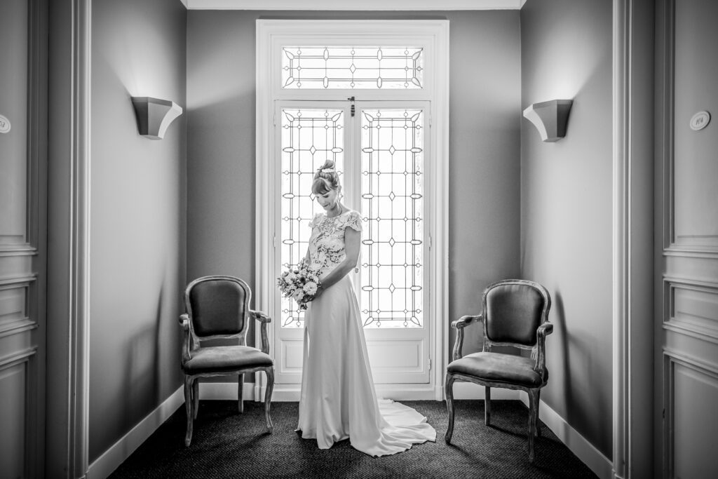 Photographie de mariage de Mathilde et Kévin. Kévin en tenue de cérémonie marron et Mathilde avec sa belle robe blanche. Ils sont debout dans l'église et Mathilde passe l'alliance au doigt de Kévin, tandis que lui la regarde avec amour . Photo en couleur. Photographie prise par un photographe de mariage professionnel à Béziers