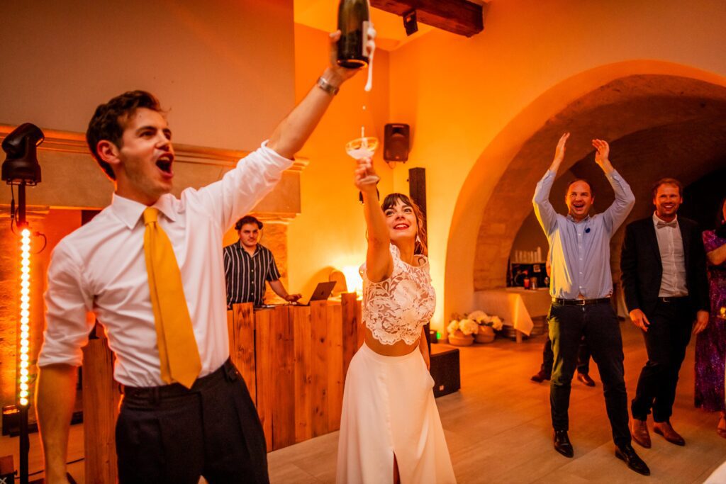 photographie de mariage de Mélanie et Alban, les mariées se promène dans la rue mains dans la mains, photographie en couleur , le marié à ouvert la bouteille de champagne qui coule dans le verre de la mariée qui est en dessosus, dans le gard