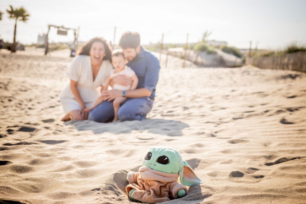 photo de famille à Montpellier en bord de plage. Les parents et l'enfant sont loin, la mise au point est faite sur Grokku qui est le doudou du petit, avec les parents en fond flou et le doudou net en première partie de photo